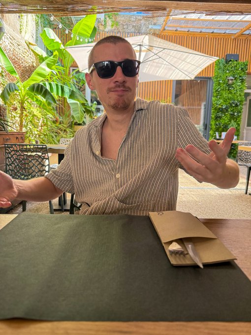 A man wearing sunglasses and a tan striped short-sleeve shirt sits at a table outdoors under a pergola, gesturing with his hands, with a green menu and a paper-wrapped utensil set in front of him.