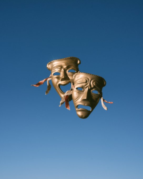 Two golden theatrical comedy and tragedy masks (joy and sorrow) appear to float against a clear blue sky, with decorative ribbons attached to their sides.