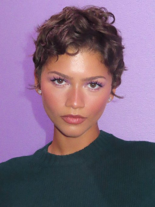 A close-up portrait of a young woman with short curly brown hair, wearing pink eyeshadow and a dark green sweater, posed against a purple background.