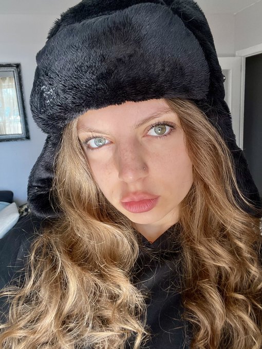 A woman with long wavy brown hair and a small nose piercing wears a fluffy black trapper hat while looking directly at the camera in a selfie. The cozy, intimate portrait complements the warm, personal tone of the post vouching for someone in the crypto community.