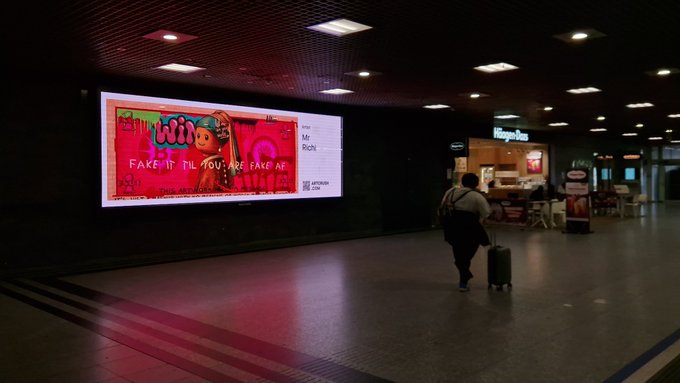 A dimly lit airport terminal where a person with a backpack pulls a suitcase past a large illuminated advertisement reading 'Lil Wiin Make it til you are fake af' by Dubai-based artist Rishi, with a Häagen-Dazs shop visible in the background.