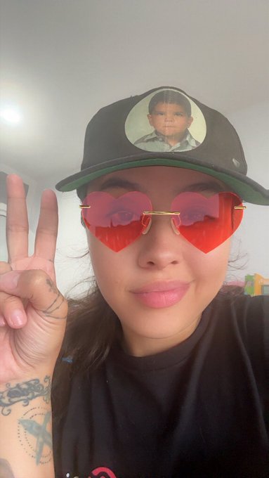 A selfie of a woman wearing red heart-shaped sunglasses and a black cap printed with a photo of a man's face, making a peace sign with a tattooed hand.