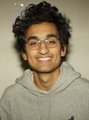 A young man with curly hair wearing glasses and a grey hoodie smiles in a simple portrait photo.