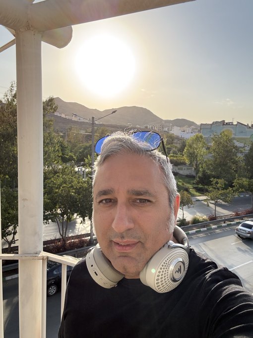 A selfie of a man with sunglasses on his head and white headphones around his neck, standing on a balcony with a view of a residential neighborhood, trees, and mountains under a bright sun.