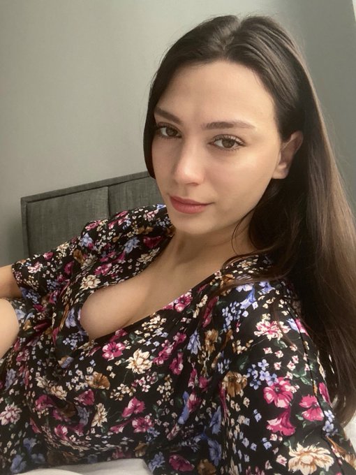 A young woman with long dark hair takes a selfie while wearing a black floral-patterned top, posing indoors against a light wall. The casual, personal photo style fits the informal weekend greeting vibe of the crypto trading update about depositing USDC and picking up the $ANSEM token on BullpenFi.