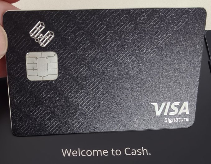 A close-up of a person's hand holding a textured black 'VISA Signature' credit card above a dark surface that reads 'Welcome to Cash.'