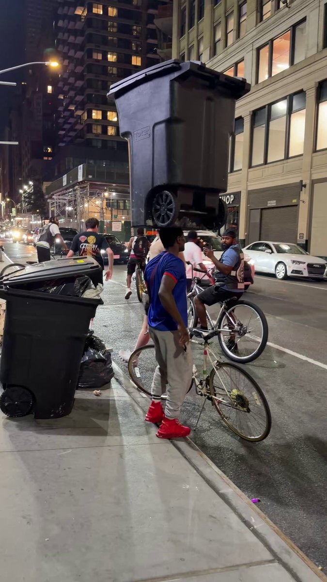 The image shows a man standing next to two trash cans, one of which has a large black bin on top of it. The man appears to be wearing red shoes and holding a bicycle with a basket attached to its back. In the background, there are other people walking around in the city street at night. A car is also visible parked nearby. The image seems to be related to an urban scene or event where the man is balancing a large trash can on his head as part of some performance or challenge.