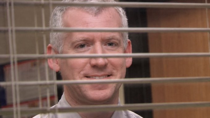 A man with gray hair smiles slightly while looking through the horizontal metal bars of what appears to be an enclosure or window blind.