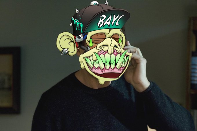 A person wearing a black sweater has a cartoonish, monstrous mask covering their face, featuring large yellow teeth and a hat labeled "BAYC".