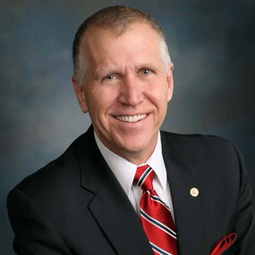 A professional headshot of a smiling older man in a dark suit with a red striped tie against a blue background.