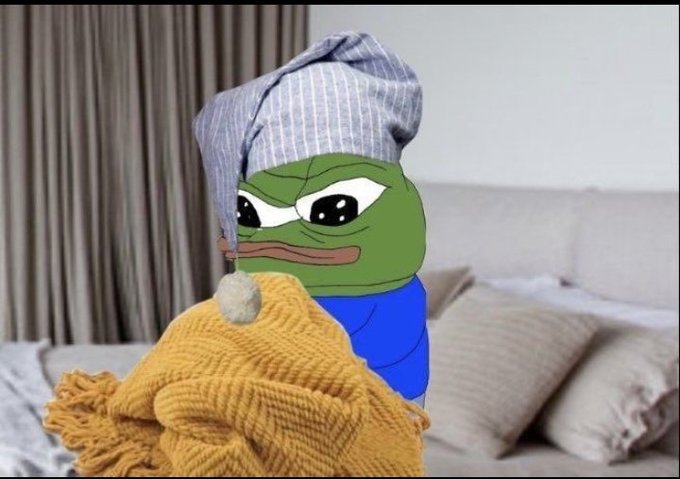 A Pepe the Frog meme edited onto a photo of a bedroom, with the frog wearing a striped nightcap and wrapped in a yellow knit blanket while sitting on a bed.