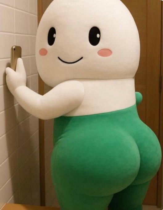 A cute mascot character with a big smiley face is pictured holding a phone against a tiled wall, presumably taking a selfie to show off its green pants. The image humorously plays on the "Made ya look" caption to promote buying the token referenced in the post.