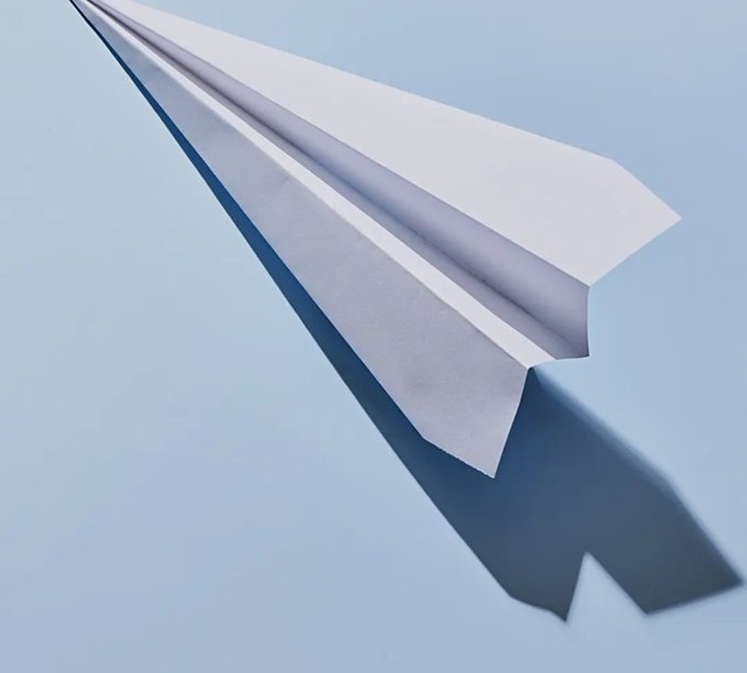 A white paper airplane soars against a clear light blue sky, casting a distinct shadow below it.