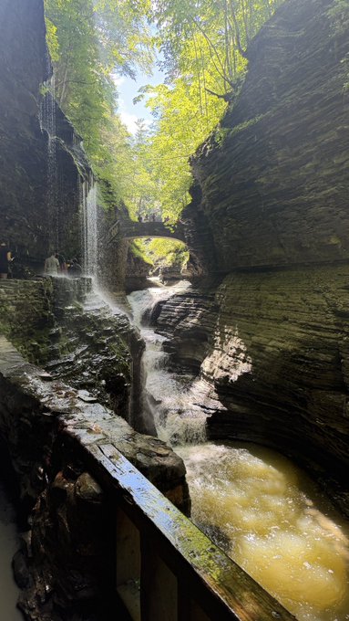 The image shows a waterfall cascading down into a river, surrounded by trees and rocks. The waterfall is located at the bottom of a steep cliff or canyon, with a bridge spanning across the water below. A person can be seen standing on top of the bridge in the background. The photo captures the beauty of nature and the serenity it offers to those who take the time to appreciate its splendor.
