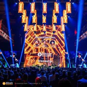 Stage photograph from the bitcoin MENA 2025 conference (Abu Dhabi, November 5-6) showing a large crowd watching a stage with orange geometric screens and speakers presenting at the Quara Factory event.