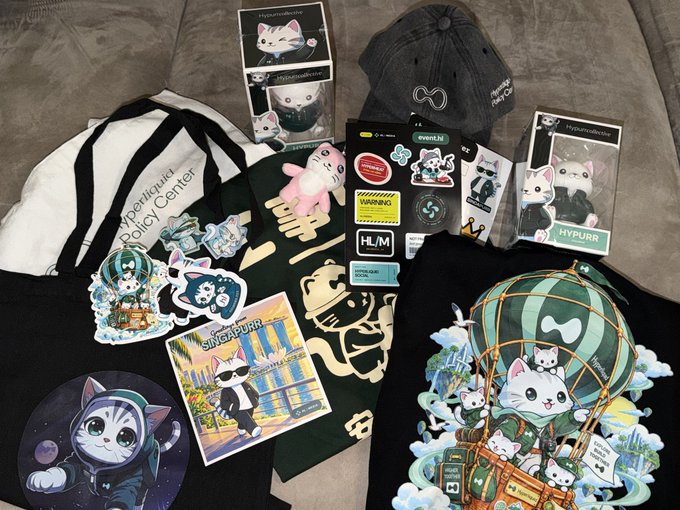 A flat lay of Hypurr merchandise and accessories, including a black cap, tote bag, hoodie, decals, stickers, boxed figures, cards, and cat-themed artwork.