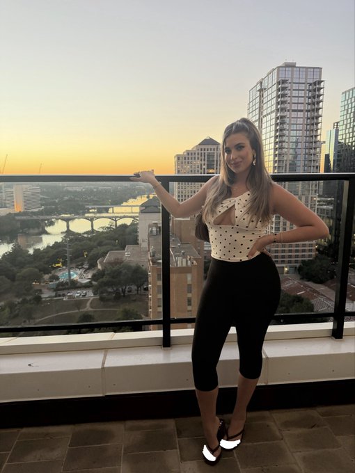 A woman poses confidently on a rooftop balcony at sunset, wearing a polka-dot one-shoulder top and black leggings, with a scenic city skyline and river visible in the background—perfectly capturing a relaxed "gm" (good morning) vibe for the start of the day.