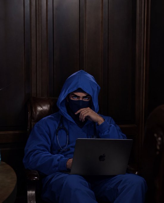 A person in a blue hooded jacket and matching face mask sits in a dark room with an Apple laptop and a stethoscope around their neck.
