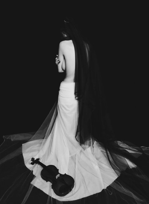 A black and white artistic photograph of a woman with long dark hair wearing a flowing white dress, sitting with a violin on the train of her dress against a black background.