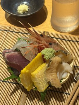 A Japanese sashimi platter featuring tuna, shrimp, tamagoyaki (egg), and other seafood arranged on a bamboo mat, served with wasabi, soy sauce, and sake.