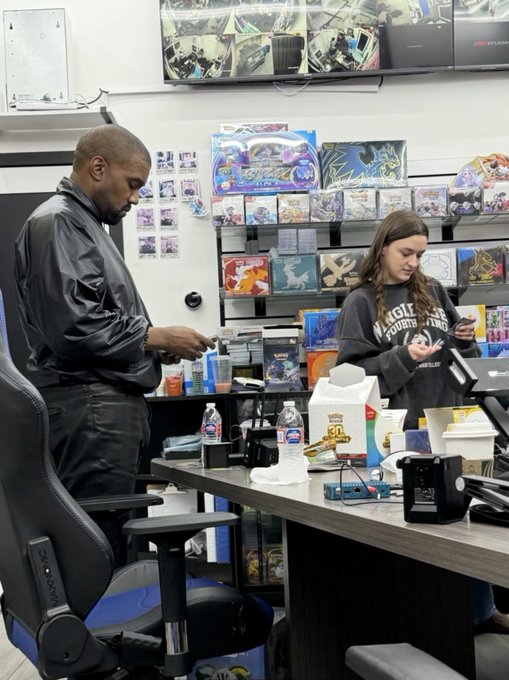 Two customers browsing inside a Pokémon trading card shop, with display cases full of card packs and boxes visible behind the counter.