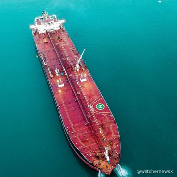 An aerial drone shot of a massive red-hulled cargo ship sailing through turquoise ocean water, with a helicopter landing pad marked 'H' visible on its deck.