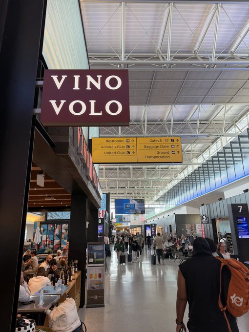 A traveler with an orange YG3 backpack walks through Austin-Bergstrom International Airport, passing the Vino Volo wine bar and directional signs pointing to gates B8-37, restrooms, Admirals/United Club, baggage claim, and ground transportation. The "Austin I am inside of you!" post reflects the excitement of actually being inside the airport after a long journey, capturing the moment of arrival in the city.
