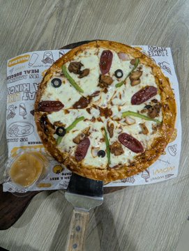 An overhead view of a small pepperoni, olive, and green pepper pizza in a branded cardboard box, accompanied by a small cup of dipping sauce and a wooden-handled serving spatula on a wood-grain table.
