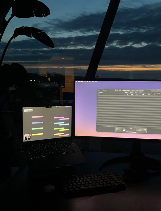 A dimly lit desk setup featuring a laptop displaying colorful horizontal bars on screen, alongside a larger monitor showing what appears to be a data interface, set against a window with a sunset cityscape view.