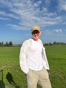 A man in a baseball cap, sunglasses, white shirt, and plaid jacket stands smiling in a lush green field under a blue sky, projecting a confident, accomplished vibe that matches the post's list of personal and professional achievements.