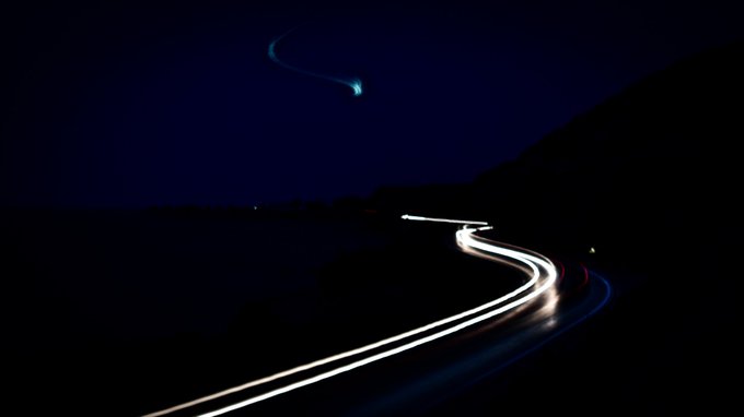 A long-exposure night photograph captures curving streaks of white and red vehicle lights winding along a dark road, with a glowing teal object leaving a wispy trail high in the night sky above. The image evokes the dynamic motion of "Linea Motus" (Line of Motion), visually translating the artwork's exploration of light, movement, and trajectory into a serene, otherworldly scene that complements the meditative digital piece.