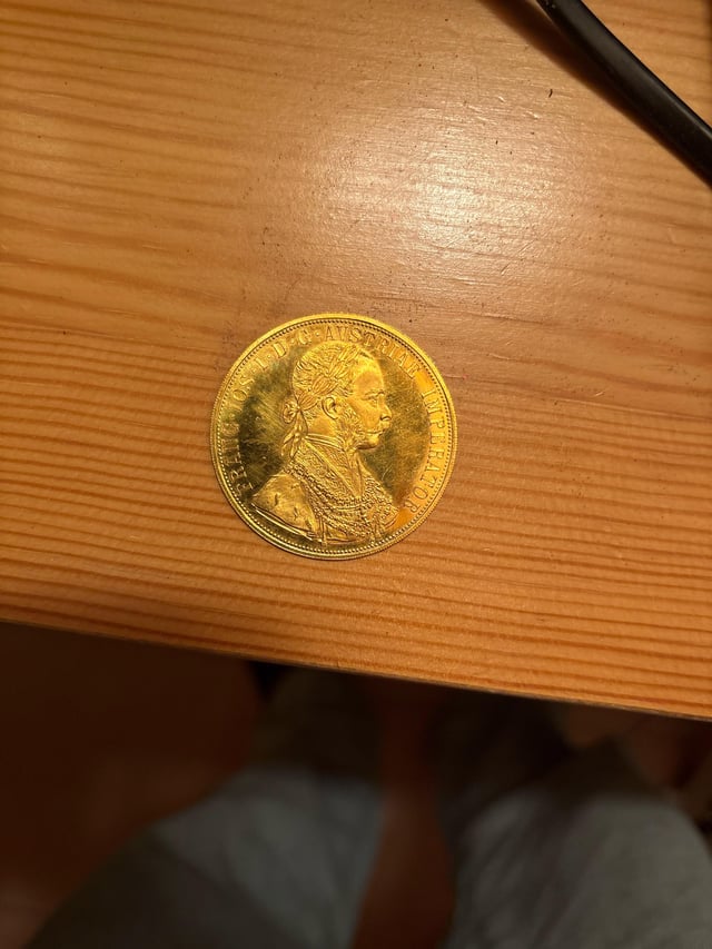 A gold-colored commemorative coin featuring a profile portrait of a man and the text "FRANC IOSEPH I D G AVSTRIAE IMPERATOR" rests on a light wooden table.