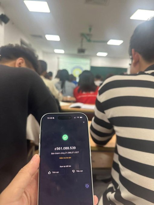 A student discreetly holds up a phone in a crowded classroom, the screen showing a successful crypto transaction of 21,088.07 USDT converted to 561,088,539 VND, perfectly capturing the post's narrative of a quiet moment of financial triumph during a lecture.