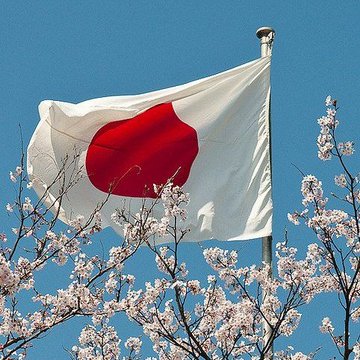 The Japanese flag waves against a clear blue sky framed by blooming cherry blossoms, symbolizing Japan's milestone legislation that officially classifies cryptocurrency as a regulated financial asset.

Key elements referenced:
- Japanese flag (red circle on white background) representing the country behind the new legislation
- Cherry blossoms (sakura) symbolizing Japan and a new beginning/era
- Date reference: early April timing aligns with Japan's fiscal year and cherry blossom season