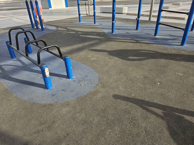 An empty outdoor playground area shows blue metal exercise equipment and bars casting long shadows on a rubberized surface.