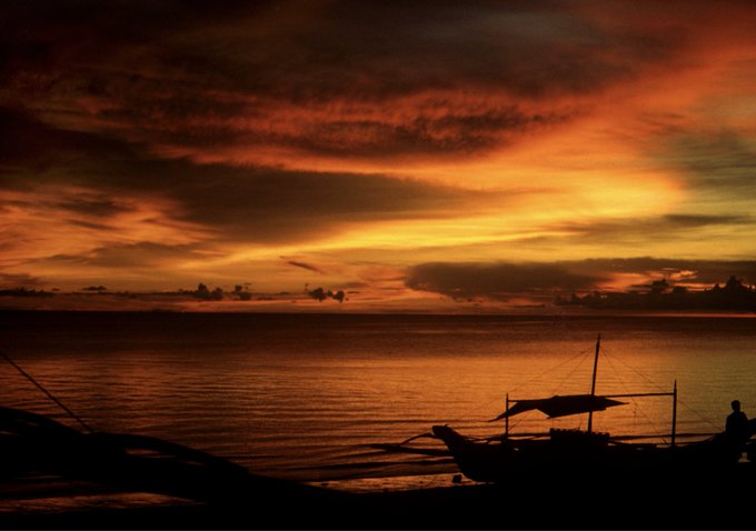 A dramatic sunset over the ocean with vibrant orange and red clouds, silhouetting a small boat with a person on the shore.