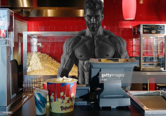 A muscular figure stands behind a movie theater concession counter, positioned next to a popcorn bucket labeled "Popcorn" and a drink cup, with red tiled walls and food warmers visible in the background. The image humorously references the post's stock ticker $GIGA by depicting a giant figure, visually punning on the ticker symbol's name.