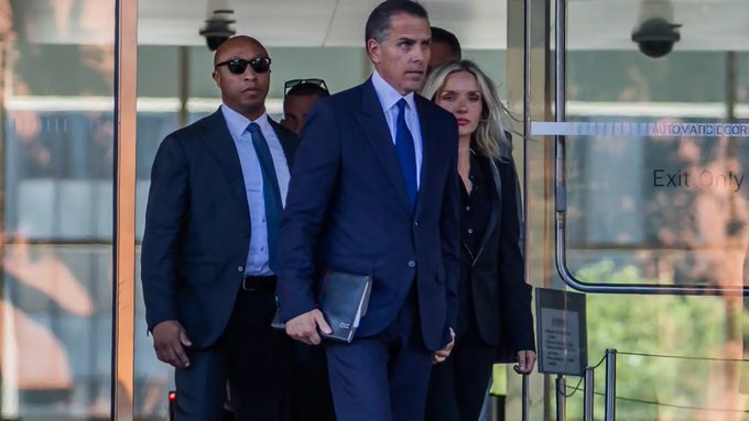 A man in a blue suit walks out of a building with 'Exit Only' on the glass door, accompanied by security personnel and a blonde woman in black.