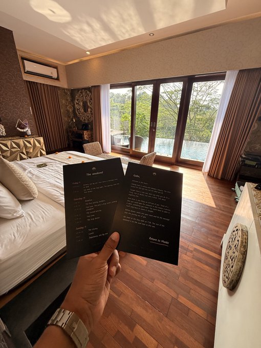 A hand holds open a menu titled "The Weekend" with sections for "Salads" and "Landing," featuring dishes like "The Garden" and "Tomato," set against the backdrop of a luxurious hotel room with a pool view, likely capturing a "GM" (Good Morning) moment during a leisurely weekend stay.