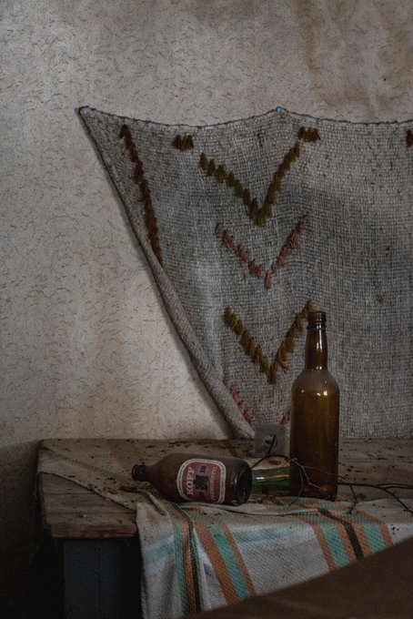 A still life photograph showing an empty brown glass bottle and a can lying on a striped cloth draped over a wooden bench, with a patterned rug hanging on the wall behind.