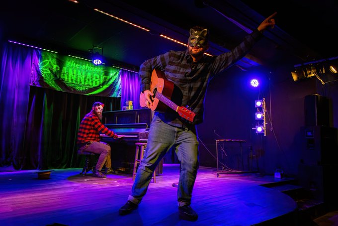 Two performers on stage at what appears to be The Green Lady Lounge; one wears a cat mask and plays guitar while the other plays piano in the background.