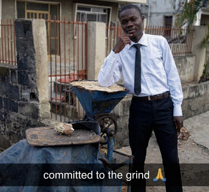 A young man in a white shirt and black tie poses next to a blue grinding machine, with the caption 'commited to the grind' and a prayer hands emoji.