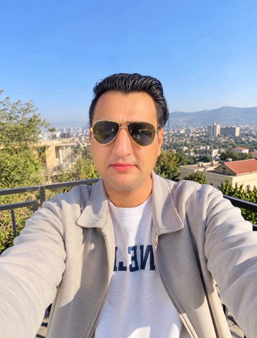 A man in aviator sunglasses and a gray jacket poses for a selfie on a sunny balcony with a cityscape and mountains in the background, his shirt displaying partial text "NE"... capturing an optimistic "Good Morning" vibe to match his wish for a prosperous new month.