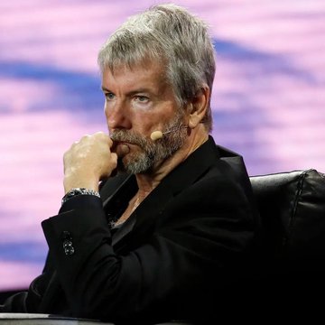 The image features a man sitting down with his hand on his chin, possibly deep in thought or contemplation. The background of the image shows a purple screen, which could be part of an event or presentation taking place at a conference room. There is also a keyboard visible in the scene, suggesting that this might be a computer-based meeting or discussion.