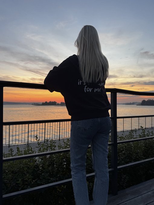 A blonde woman stands facing away from the camera, leaning on a railing and looking out at a sunset over the water, wearing a black sweatshirt with the text "it's your for me" and blue jeans.