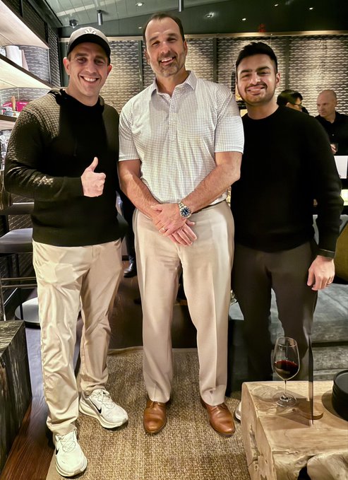Three men posing together at what appears to be a restaurant or bar; the central figure is notably tall, wearing a light shirt and khaki pants, flanked by two smiling men in casual attire.