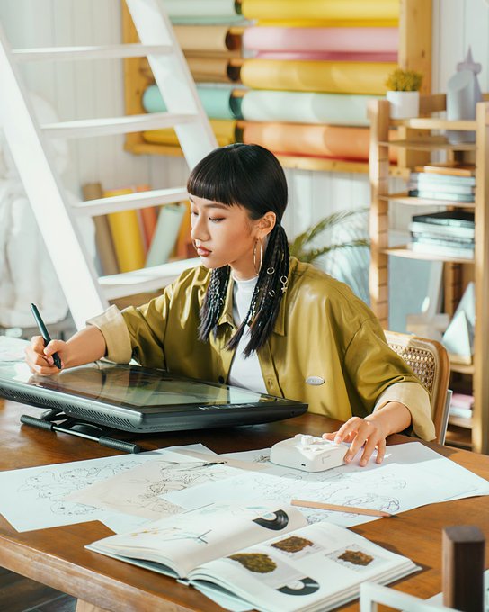 A young woman with dark hair in a green jacket sits at a wooden desk in a bright studio, drawing on a graphics tablet with sketches and an open book in front of her.