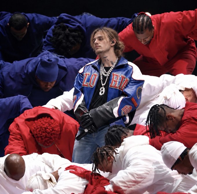 A male performer wearing a blue and white jacket stands among a group of people kneeling in red, white, and blue hooded outfits during what appears to be a stage performance.