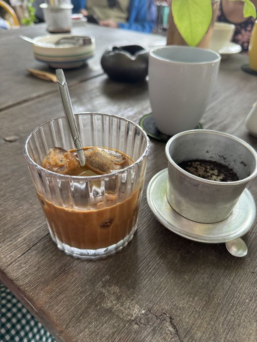 A wooden table with a glass of iced coffee, a small ceramic cup of black coffee, and a white mug in the background.
