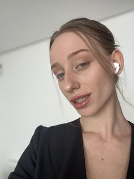 A selfie of a young woman with light brown hair pulled back, wearing white AirPods and a black top against a plain white background.
